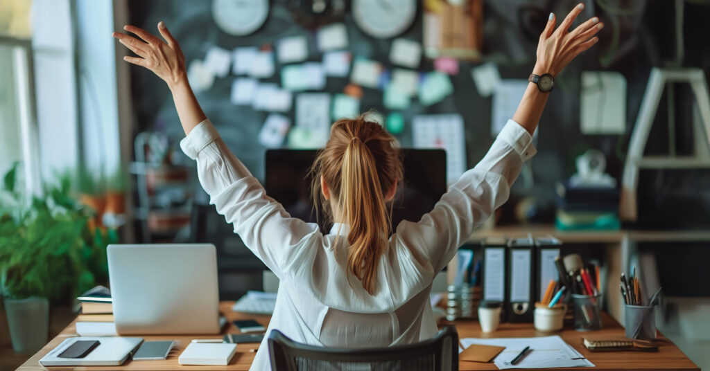Eine Frau sitzt im Büro vor ihrem Rechner und streckt freudig die Arme nach oben. Ein Zeichen für Motivation am Arbeitsplatz.