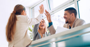 Ein glückliches Team in einem Büro-setting gibt sich High fives. Das Ergebnis von Führungskräften die ihr e Mitarbeiter fördern und motivieren