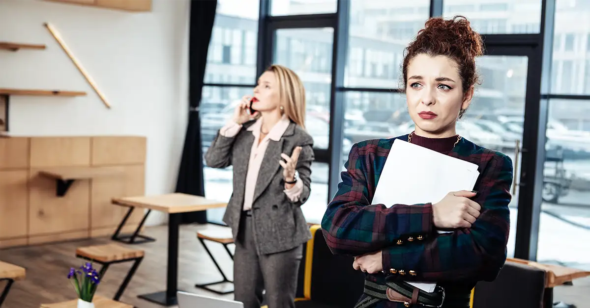 Junge, verunsicherte Mitarbeiterin mit Unterlagen im Arm steht im Vordergrund, im Hintergrund eine streng wirkende Chefin beim Telefonieren – Symbolbild für typische Führungsfehler im Mittelstand