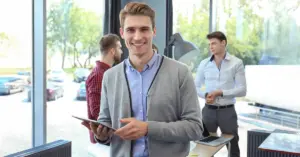 Junger Mann übernimmt eine Führungsposition ohne Führungserfahrung, lächelnd mit Tablet im Büro neben Kollegen
