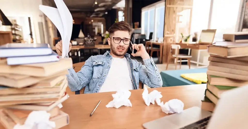 Ein überforderter Mitarbeiter telefoniert an einem vollen Schreibtisch voller Unterlagen.