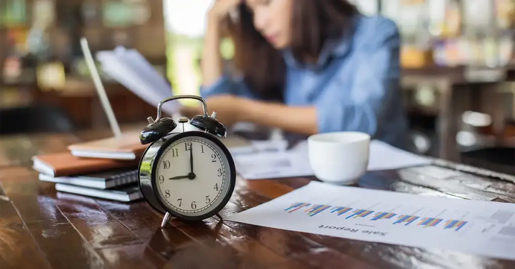 Eine Angestellte sitzt gedankenversunken an ihrem Schreibtisch. Im Vordergrund steht ein mechanischer Wecker als Symbol für die Überstunden ohne Anordnung.