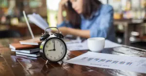 Eine Angestellte sitzt gedankenversunken an ihrem Schreibtisch. Im Vordergrund steht ein mechanischer Wecker als Symbol für die Überstunden ohne Anordnung.