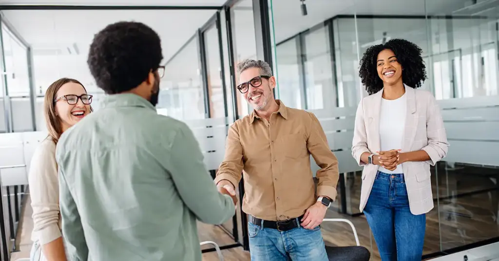 Eine Führungskraft begrüßt ein neues Teammitglied beim Onboarding neuer Mitarbeiter. Weitere Teammitglieder stehen erwartungsvoll und vorfreudig daneben und freuen sich, das neue Teammitglied willkommenzuheißen.