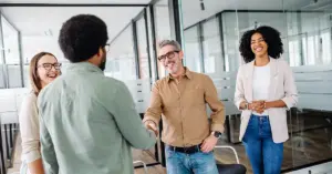 Eine Führungskraft begrüßt ein neues Teammitglied beim Onboarding neuer Mitarbeiter. Weitere Teammitglieder stehen erwartungsvoll und vorfreudig daneben und freuen sich, das neue Teammitglied willkommenzuheißen.