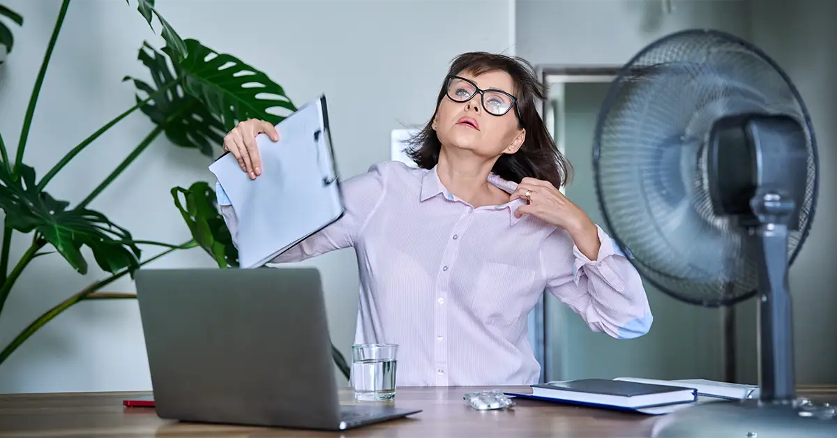 Eine Mitarbeiterin am Schreibtisch leidet unter der Hitze am Arbeitsplatz und versucht sich mit einem Ventilator und einem Clipboard Luft zuzufechern.